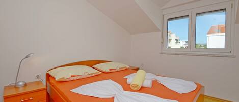 A bedroom with an orange bed, white pillows, a wooden nightstand with a lamp, and a window with a view of buildings.