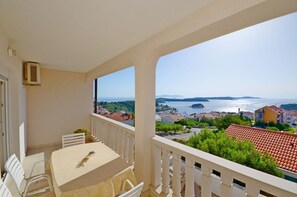 Un balcon avec une table et des chaises, donnant sur une ville côtière avec ses bâtiments et une étendue d’eau.