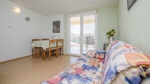A room with a sofa, a dining table with chairs, a painting on the wall, and a window with blinds. - Apartments Marija (Razanj)