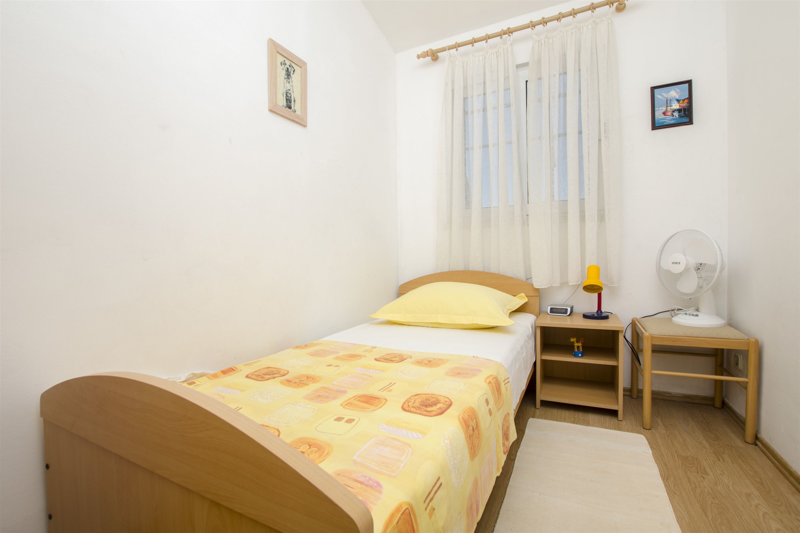 Un dormitorio pequeño y ordenado con una cama individual, una mesita de noche y un ventilador de pared.