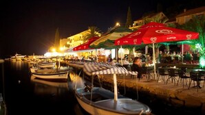 A waterfront restaurant with outdoor seating, boats docked nearby, and a building with illuminated windows in the background. - Apartments Oliva (Maslinica)