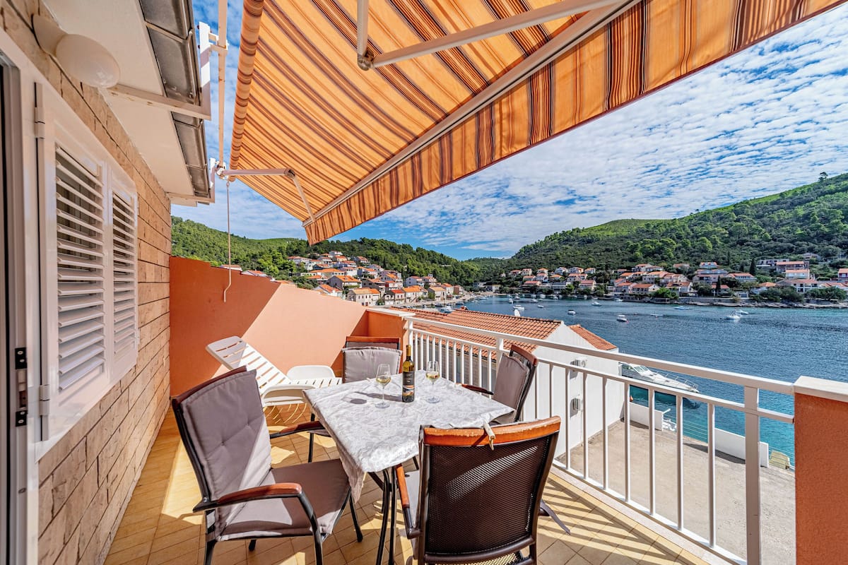 A balcony with a table and chairs, overlooking a body of water and a hillside town.