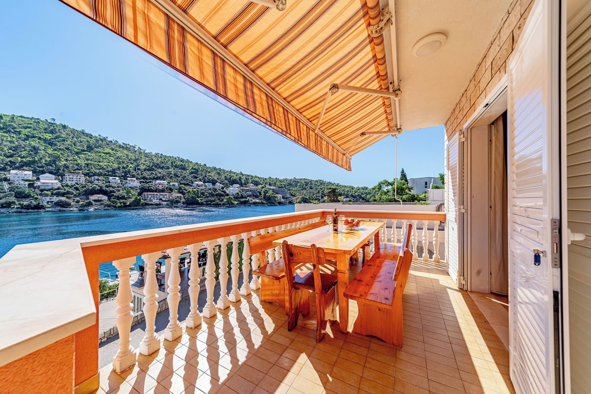 A wooden deck with a table and chairs, overlooking a body of water and a hillside.
