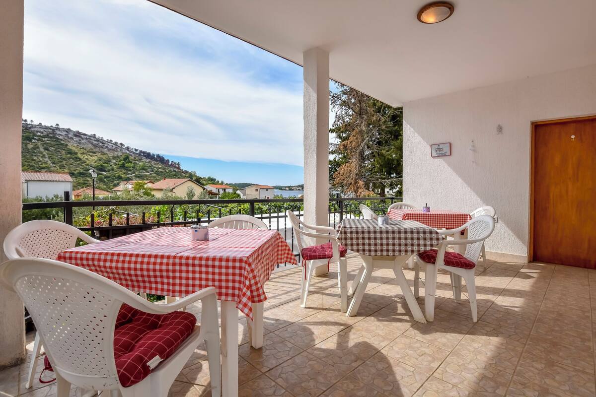 A balcony with white chairs and tables, a view of houses and a hill, and a wooden door.