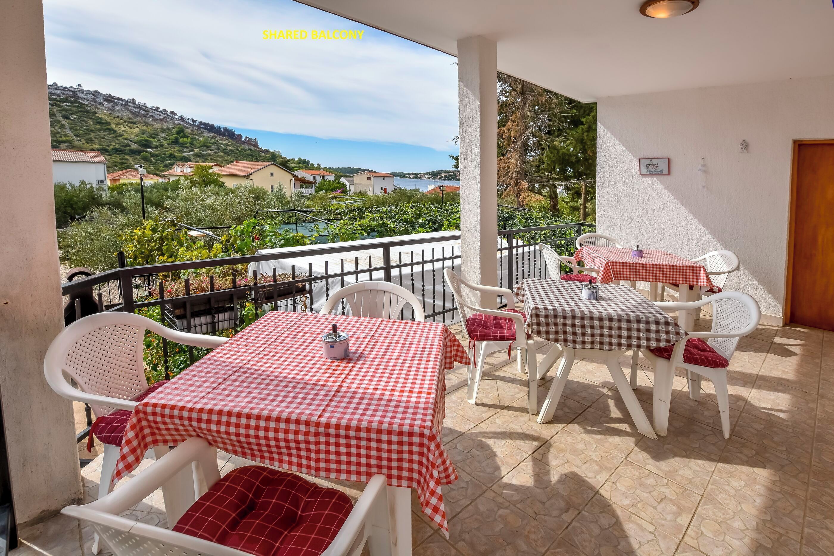 A shared balcony with white plastic chairs and tables covered with red and white checkered tablecloths.