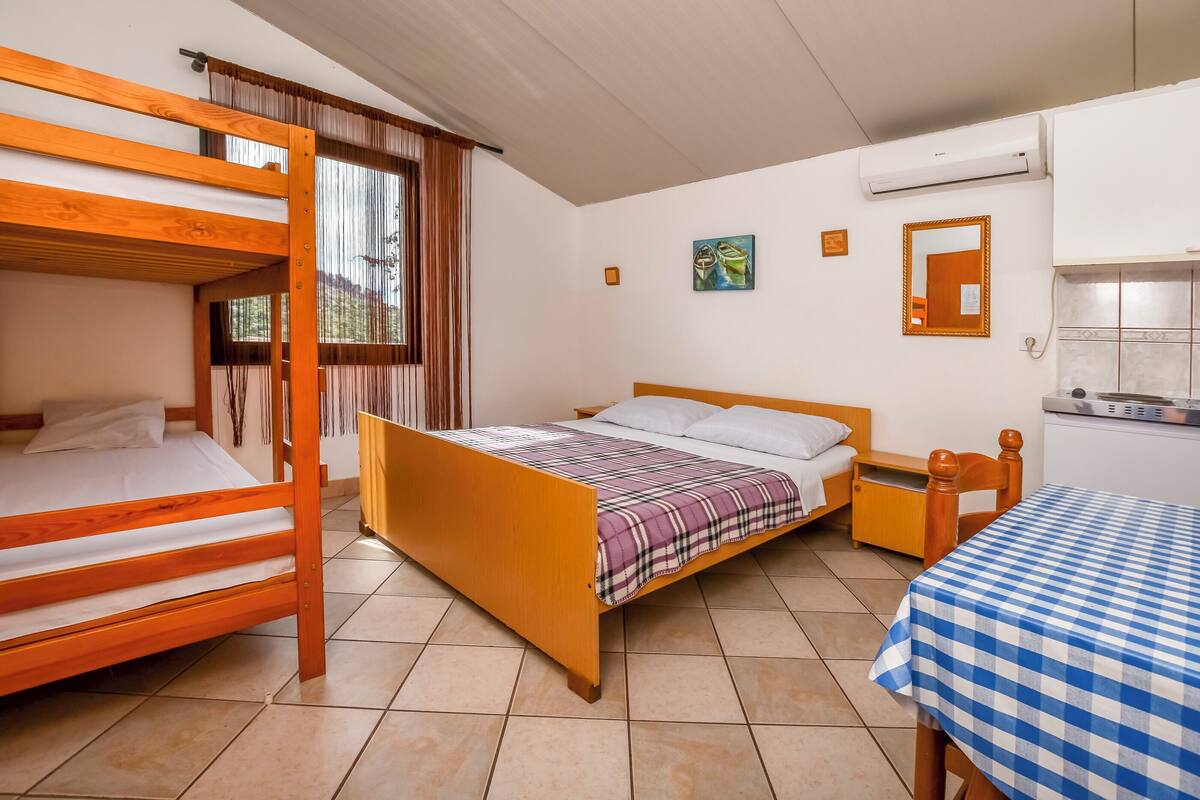 A room with a bunk bed, a single bed, a small table with a checkered tablecloth, and a window with curtains.