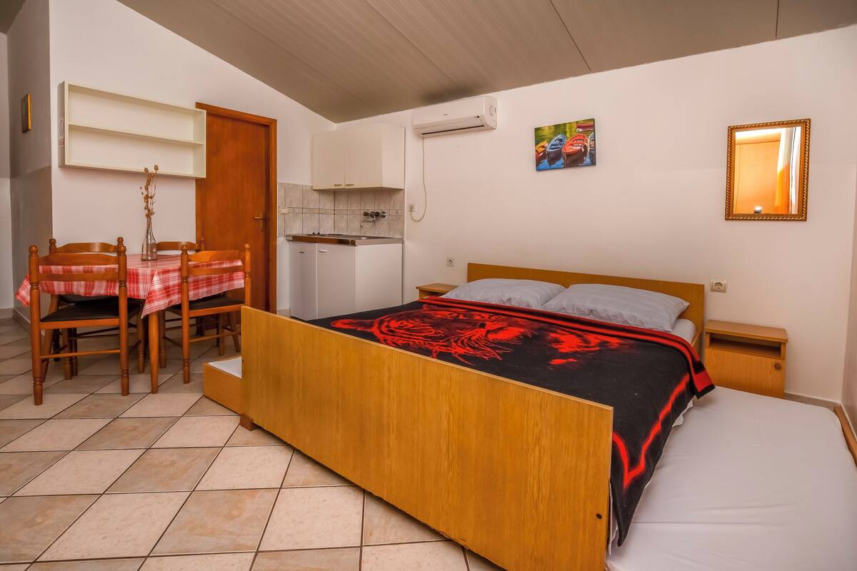 A bedroom with a wooden bed, a small bedside table, a nightstand, a red and black bedspread, a white pillow, a wooden chair, a small table with a red and white checkered tablecloth, a white door, a white wall, a white shelf, a white air conditioning unit, and a small kitchen area with a sink and cabinets.
