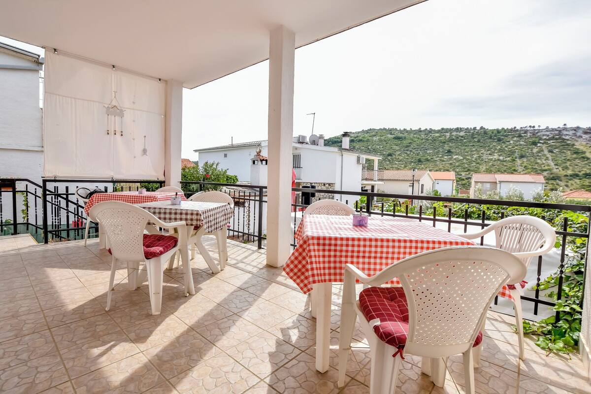 A balcony with white chairs and tables, a white awning, and a view of a hillside with buildings.
