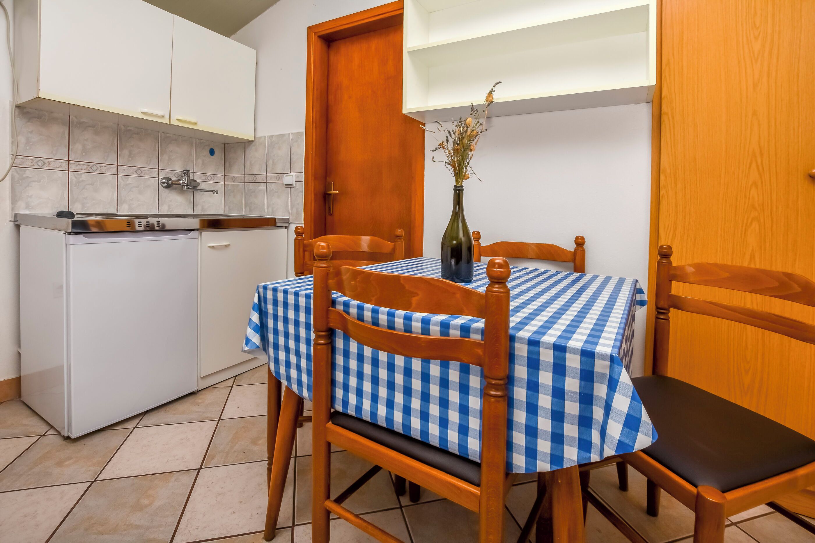 Une cuisine de taille réduite, avec un réfrigérateur, une table à manger en bois recouverte d’une nappe à carreaux, et un vase contenant des fleurs séchées placé sur la table.