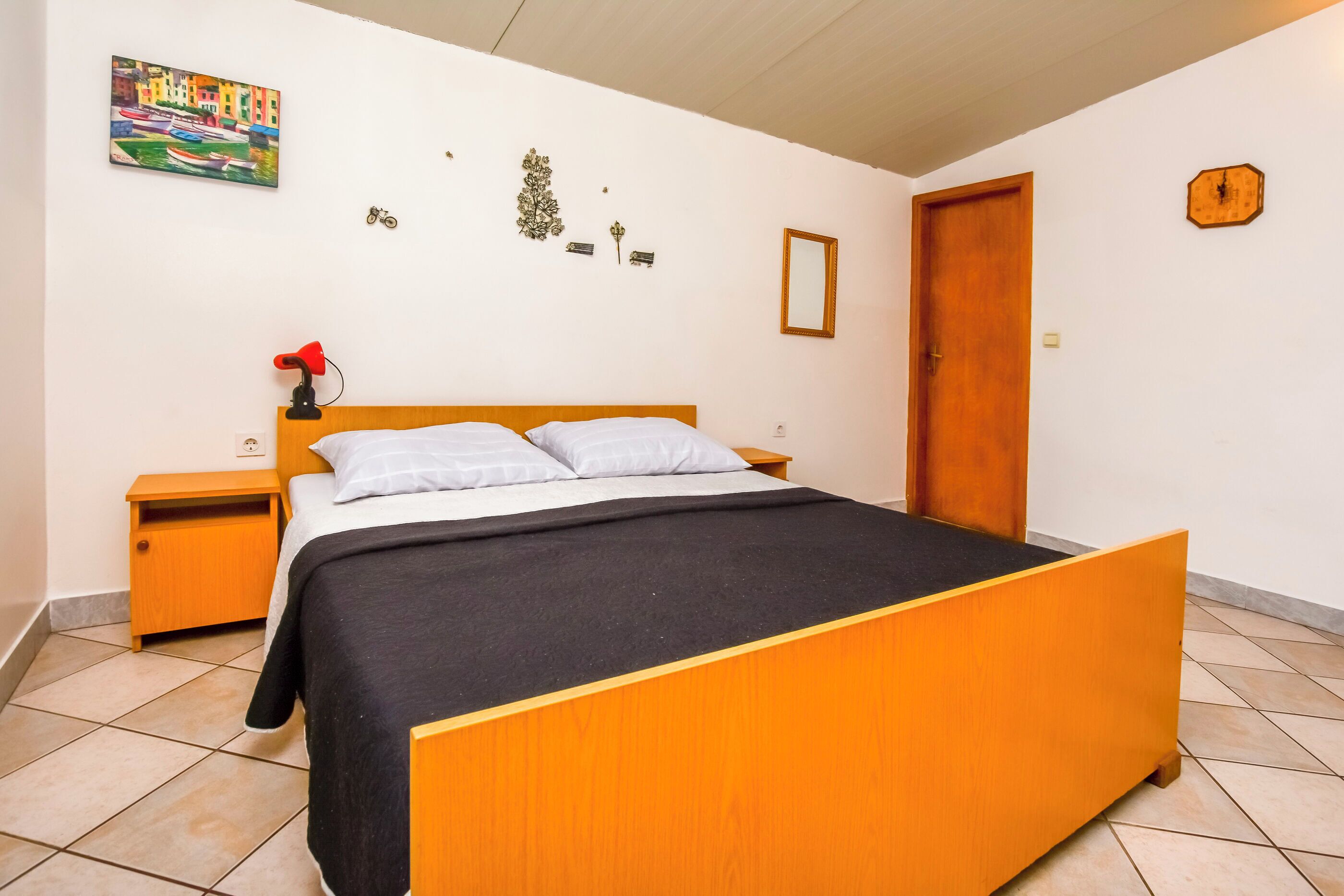 A bedroom with a wooden bed, a nightstand, a red lamp, and a painting on the wall.