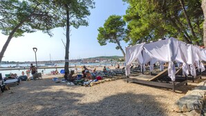 Ein Strand mit Liegestühlen, einem überdachten Sitzbereich und Blick auf das Meer und Boote im Hintergrund.