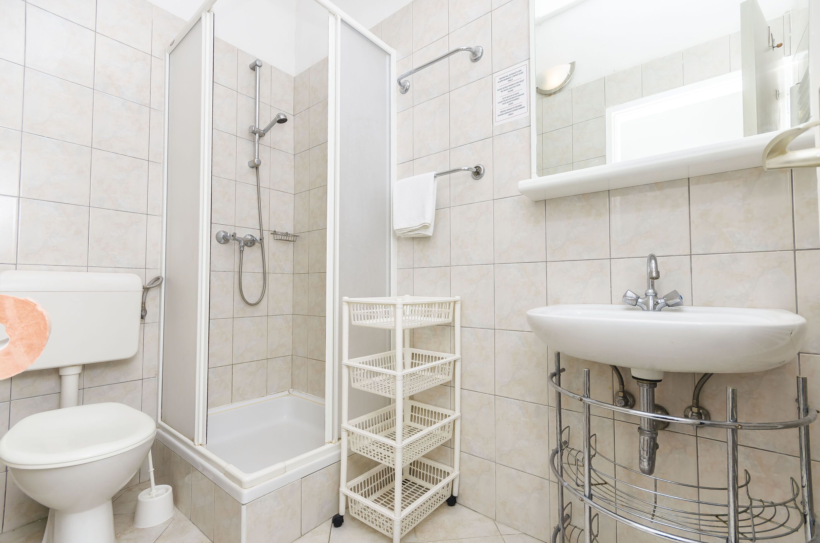 A bathroom with a toilet, shower, sink, and a white storage rack.