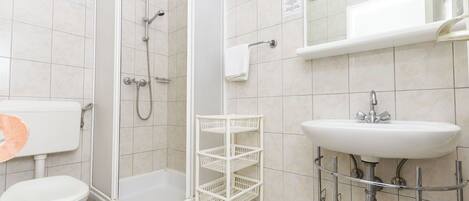 A bathroom with a toilet, shower, sink, and a white storage rack.
