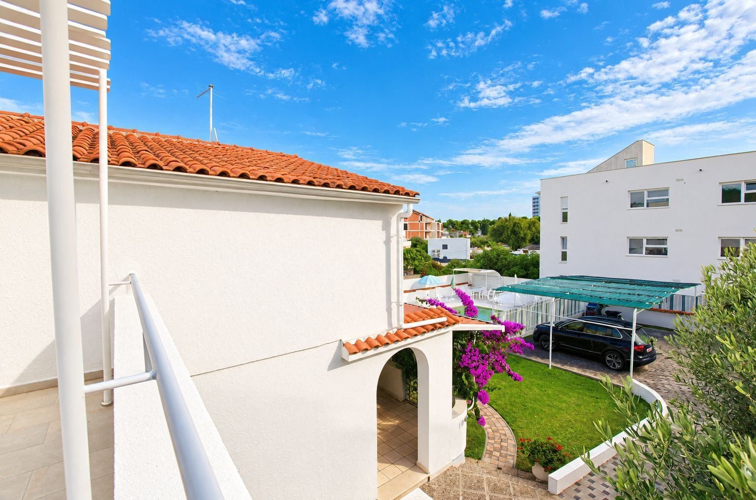 A residential building with a tiled roof, a balcony with a green awning, and a garden with purple flowers.