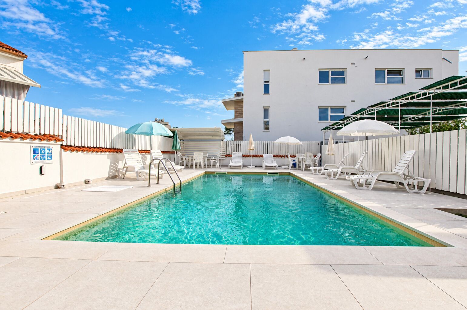 A swimming pool with sun loungers and umbrellas, surrounded by a white building with a tiled deck.