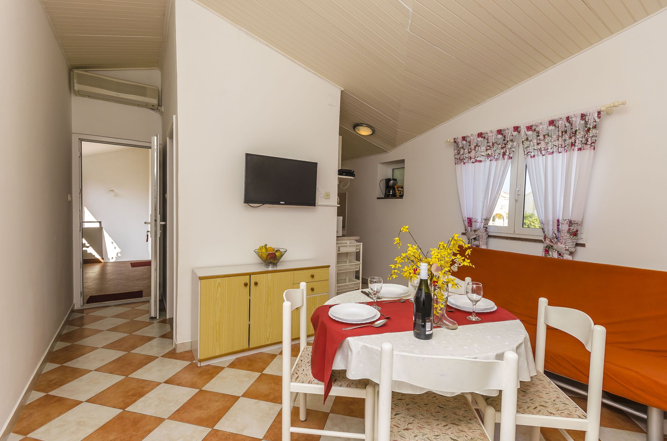 A dining area with a checkered floor, a table set with plates, a vase with yellow flowers, and a television mounted on the wall.