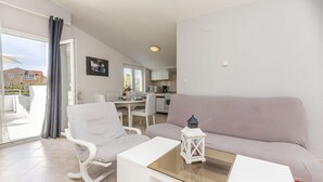Een moderne woonkamer met een bank, een witte fauteuil, een glazen salontafel en een keuken op de achtergrond.