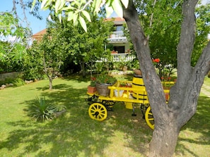 A yellow cart with potted plants parked in a lush garden with a house in the background. - Apartment Tonkica (Vodice)