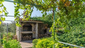 A stone-built outdoor kitchen with a brick oven, surrounded by lush greenery and a metal pergola.