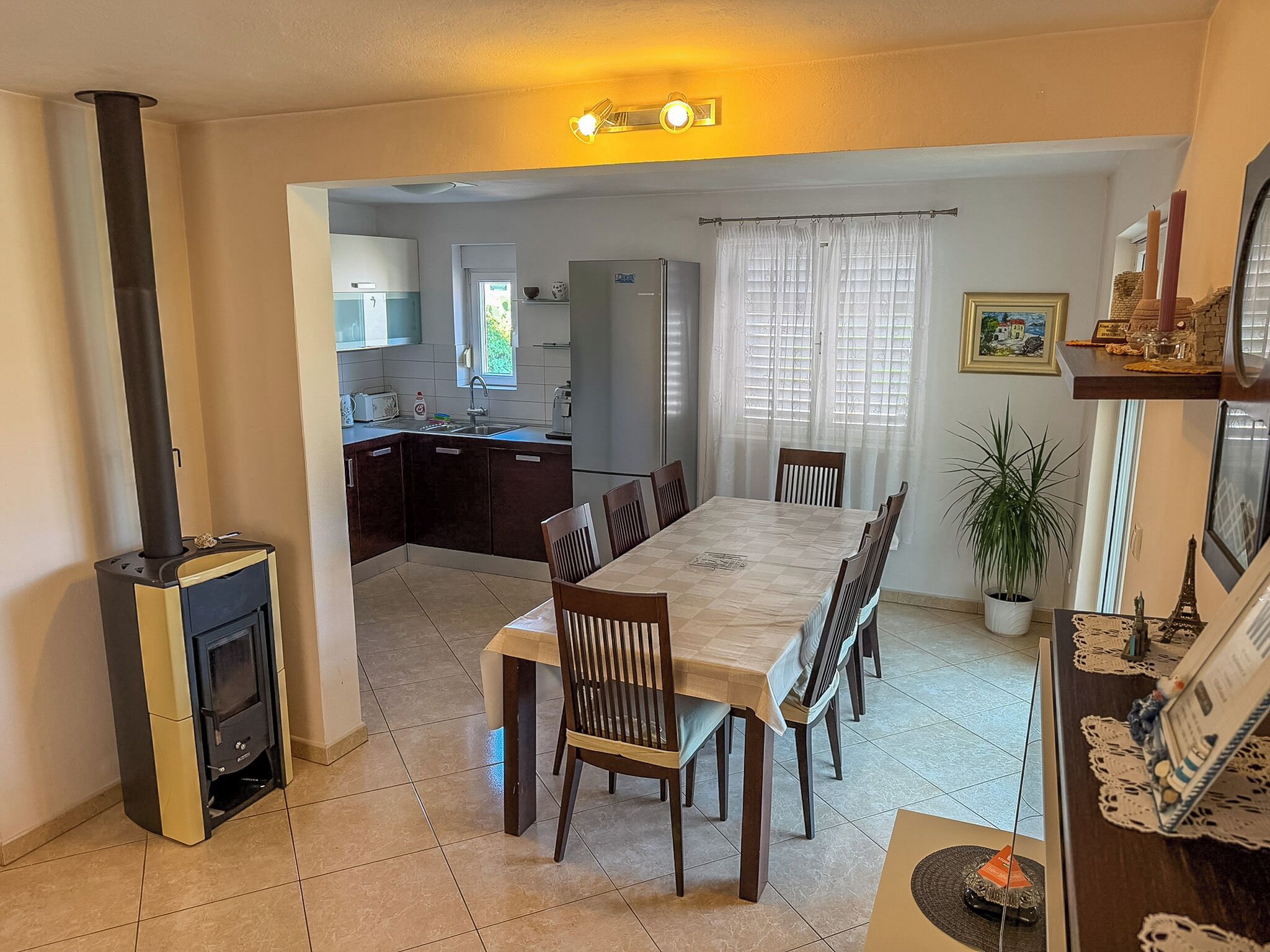 A dining area with a long table and chairs, a kitchen in the background, and a fireplace.