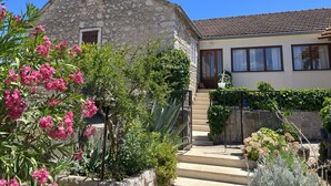 A stone house with a tiled roof, a wooden door, and a small window. There are steps leading to the door, and a garden with various plants and flowers. - House Lara (Vrboska)