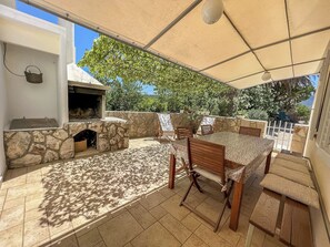An outdoor patio with a stone fireplace, a table and chairs, and a covered area with a view of trees and a clear sky. - House Lara (Vrboska)