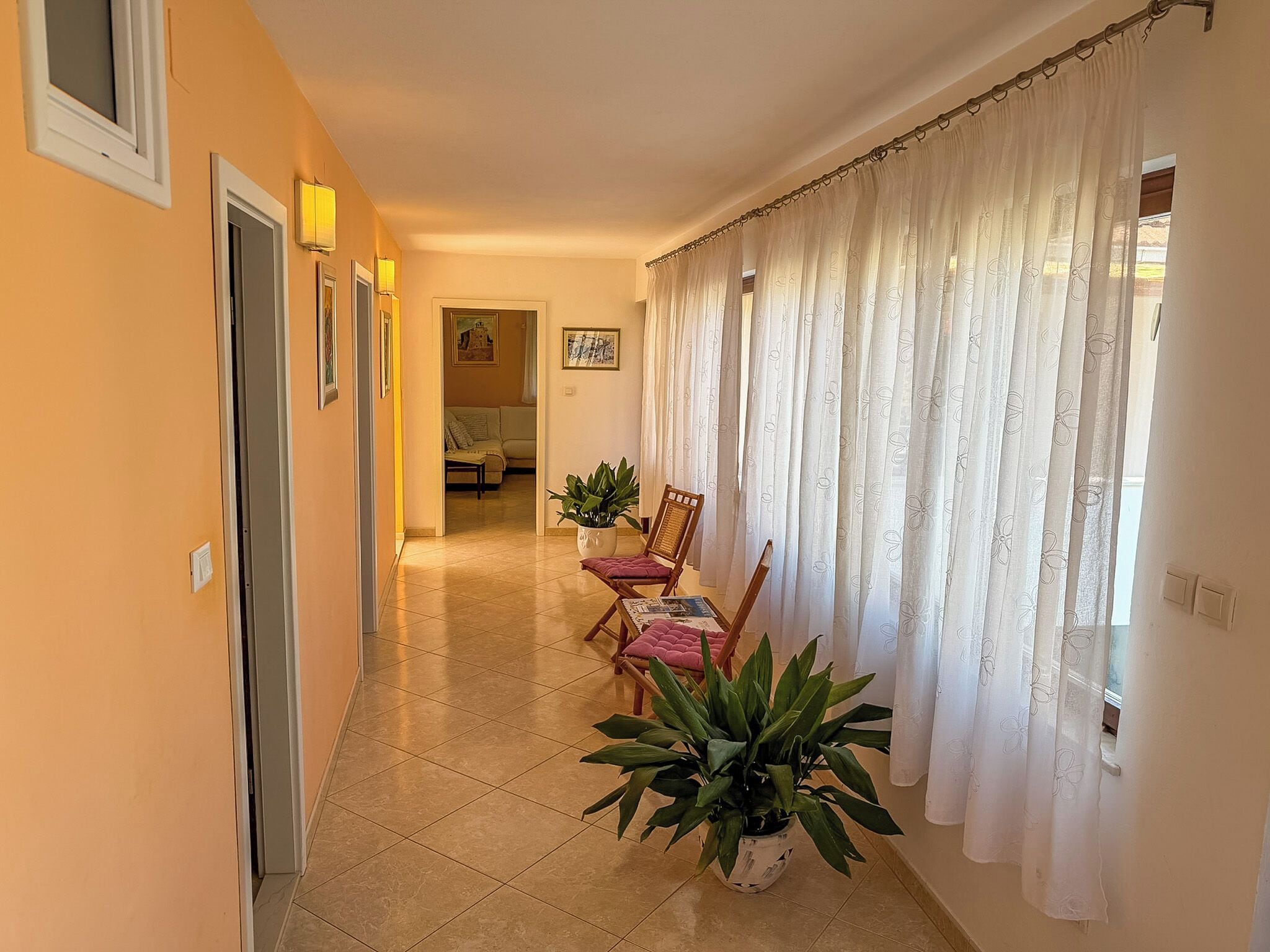 A hallway with a dining area, featuring a table with chairs, potted plants, and a view through sheer curtains.