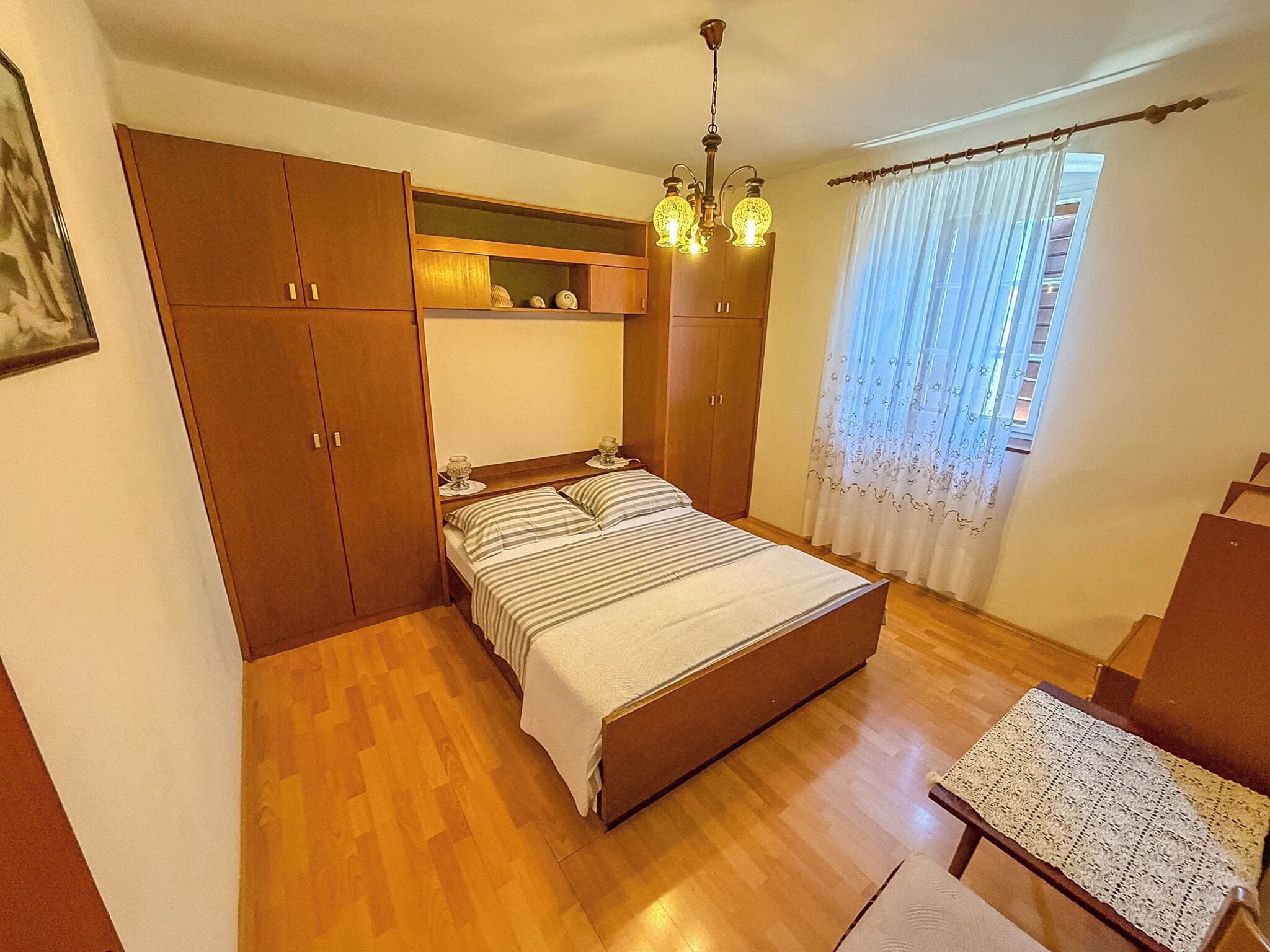 Ein Schlafzimmer mit Bett, Holzschrank, kleinem Tisch und einem Fenster mit Vorhängen.