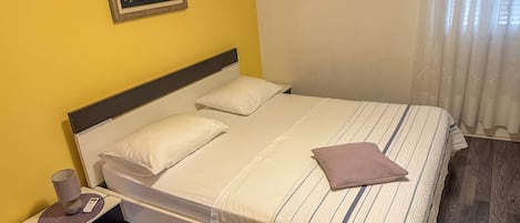 A single-bed room with a wooden floor, a bedside table with a lamp, and a painting on the wall.