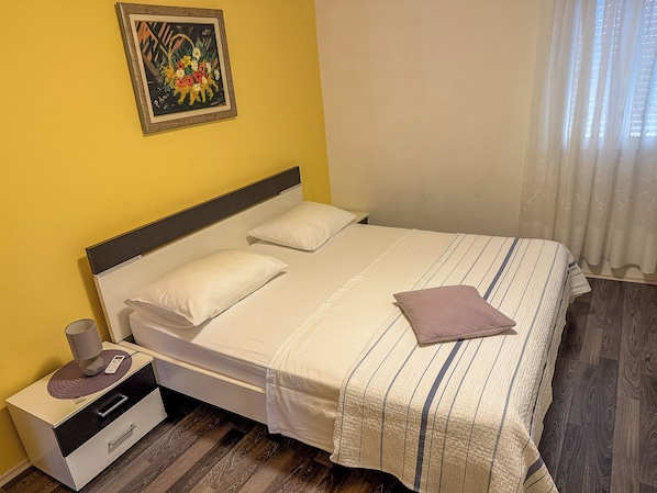 A single-bed room with a wooden floor, a bedside table with a lamp, and a painting on the wall.