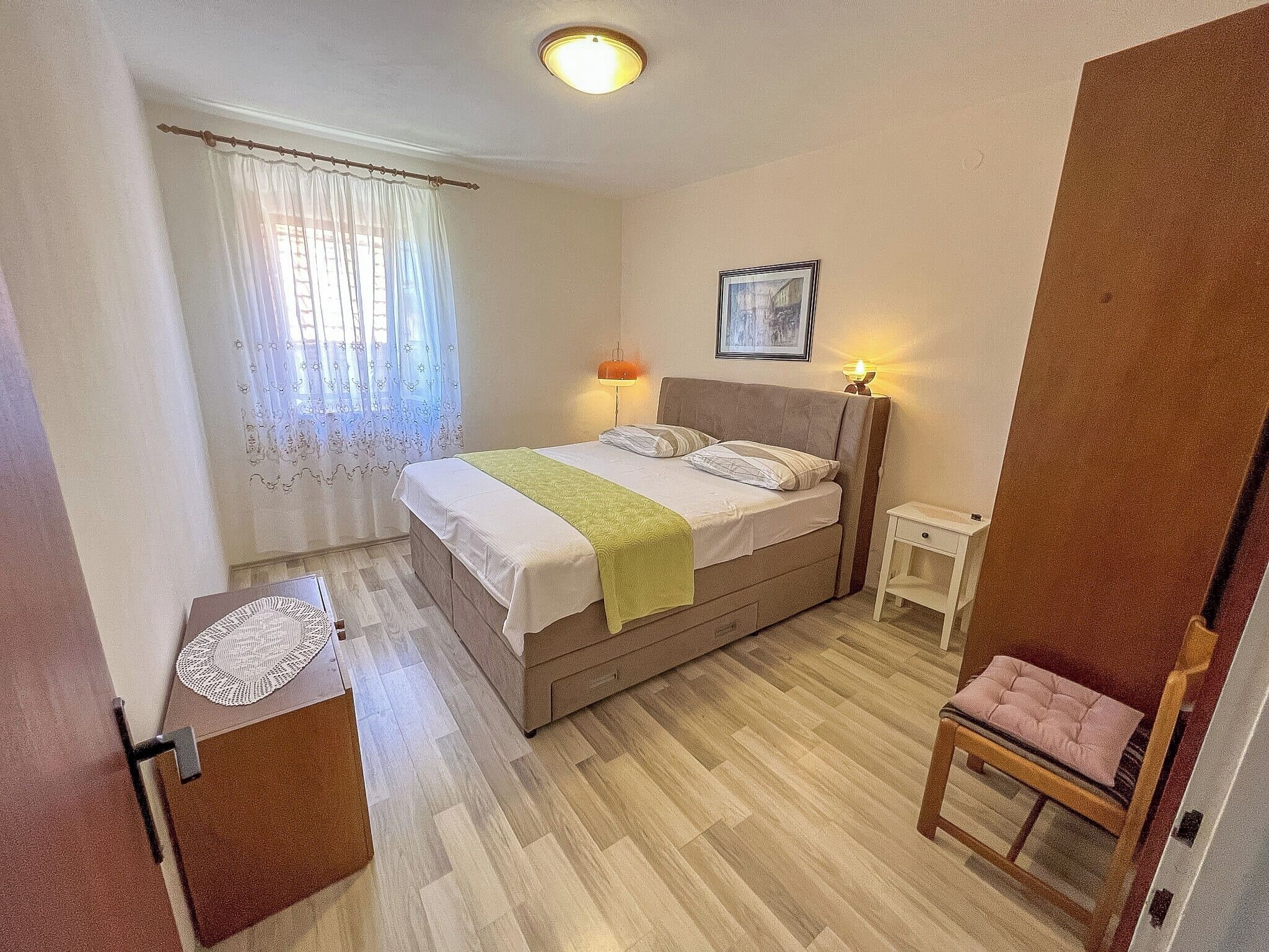 A bedroom with a bed, bedside lamps, a small table, a chair, and a picture on the wall.