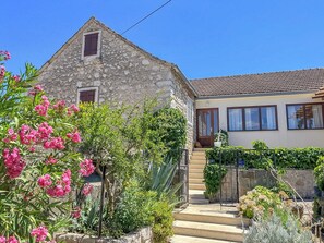Un bâtiment en pierre avec une petite fenêtre et une porte, entouré d’un jardin où poussent des fleurs roses et des plantes vertes.