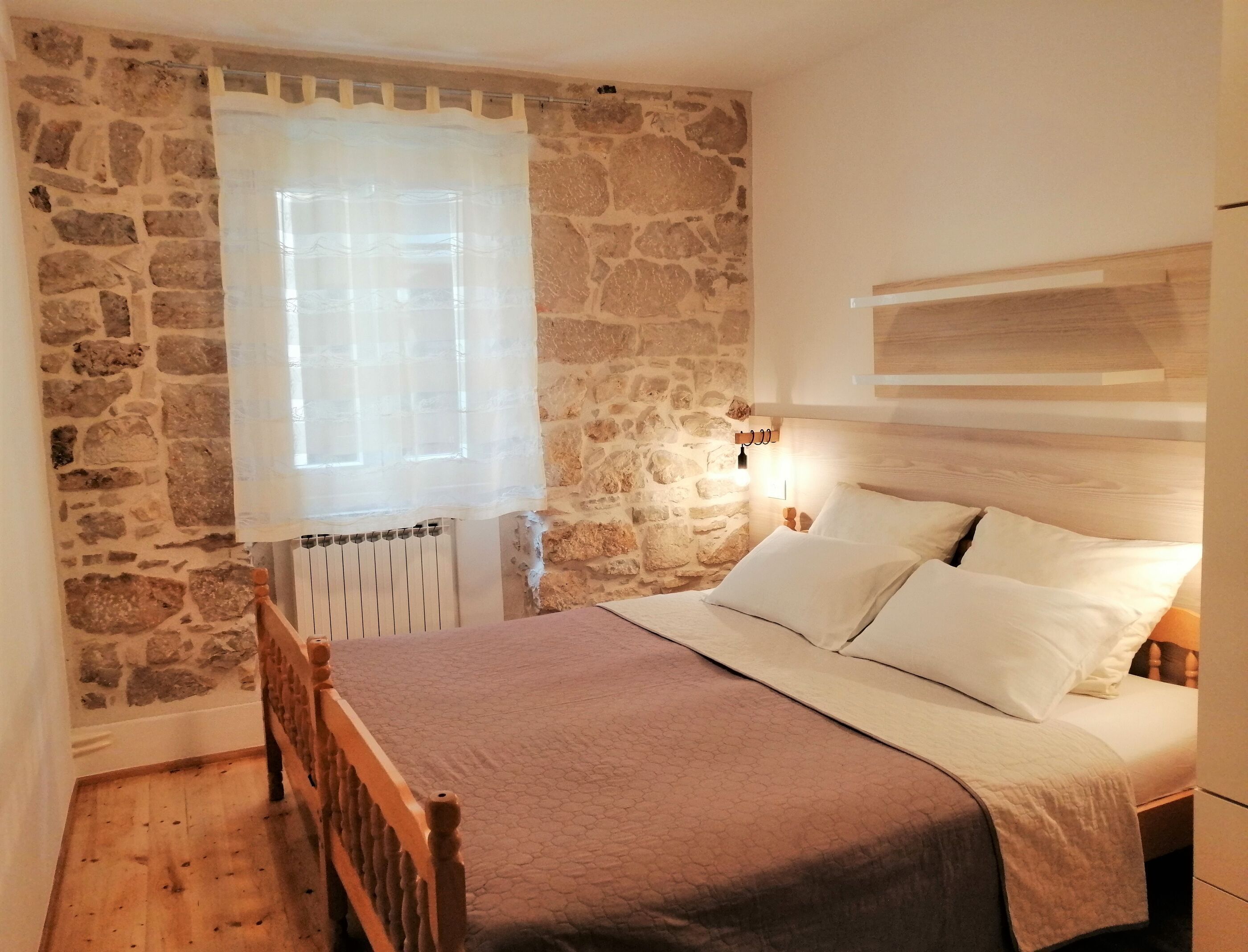 Un dormitorio con pared de piedra, estructura de cama de madera, colcha blanca y una ventana con cortina translúcida.