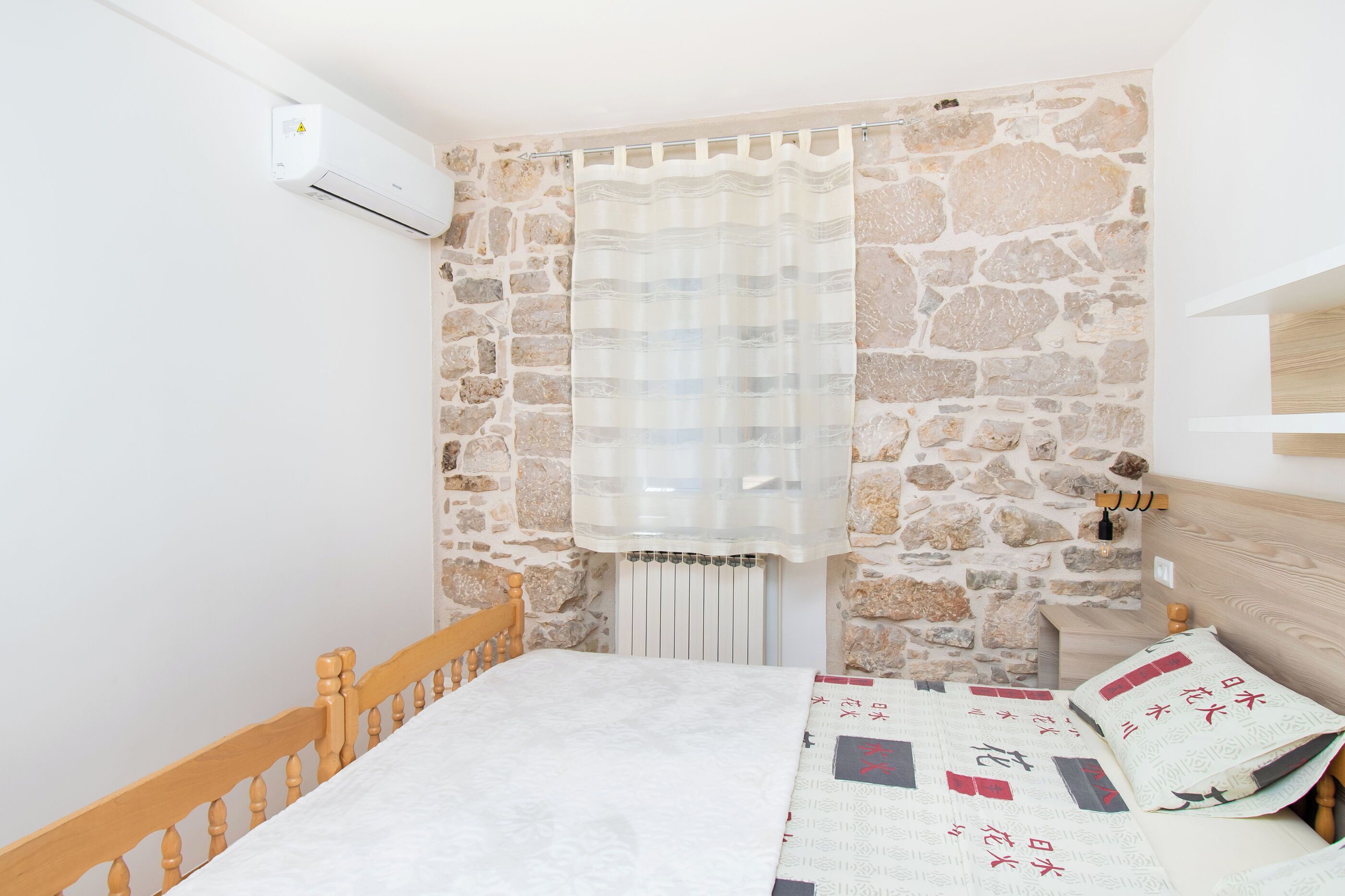A bedroom with a stone wall, a wooden bed frame, a white bedspread, and a window with a sheer curtain.