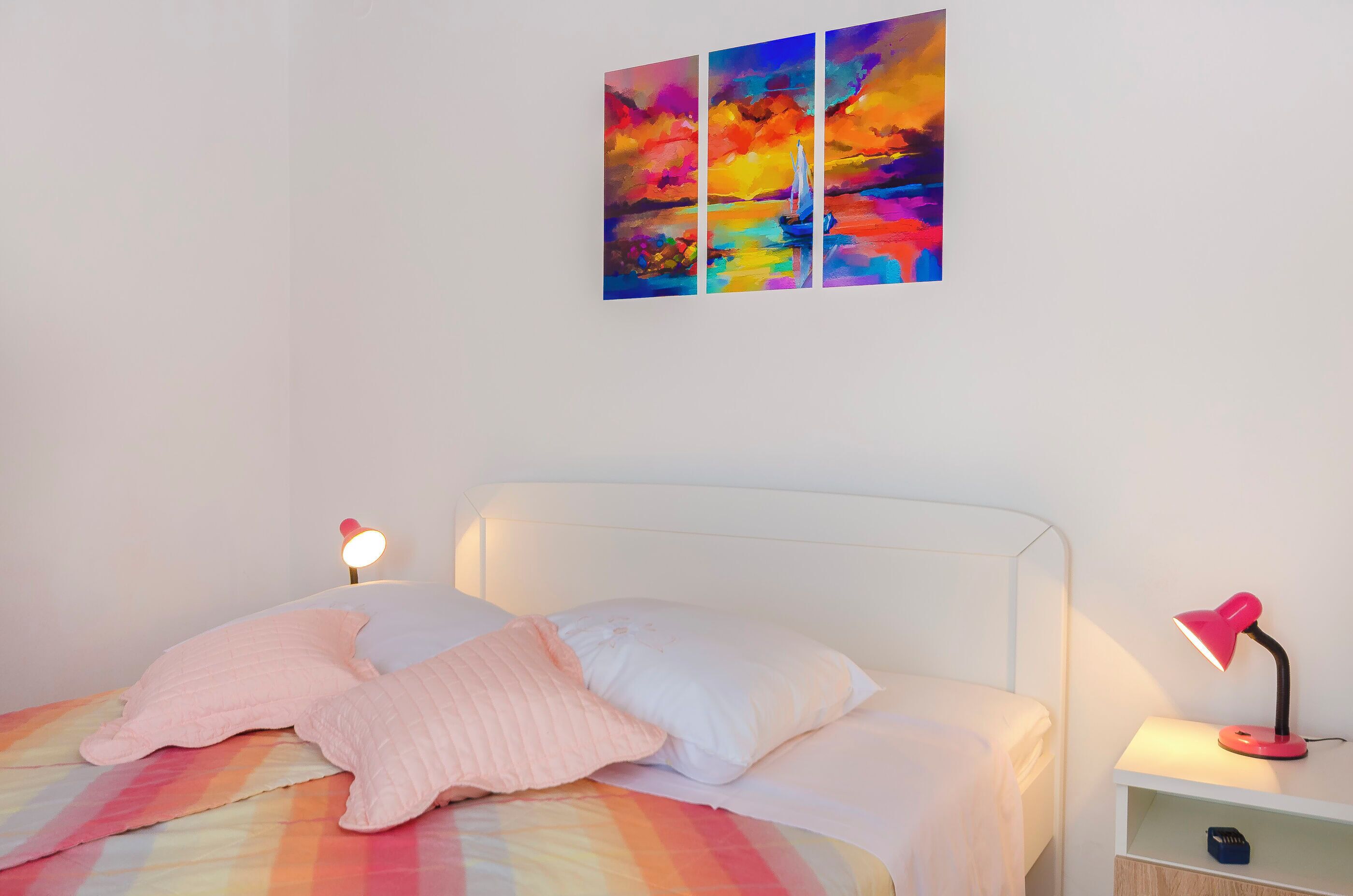 A bedroom with a bed, pillows, a bedside table with a lamp, and a wall with a colorful painting.