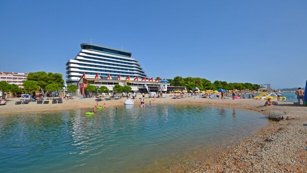 Une plage avec un hôtel moderne en arrière-plan, un ciel dégagé et des gens qui profitent de l’eau.