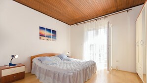 Une chambre avec un plafond en bois, un lit recouvert d’une courtepointe blanche, une table de chevet avec une lampe et une fenêtre ornée de rideaux légers.