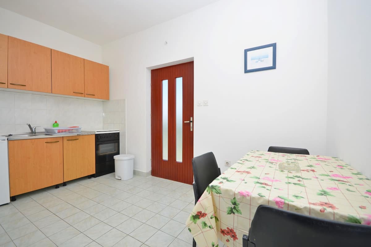 A compact kitchen with wooden cabinets, a sink, and a stove. There is a dining table with a floral tablecloth and two chairs. A white trash bin is visible near the door.