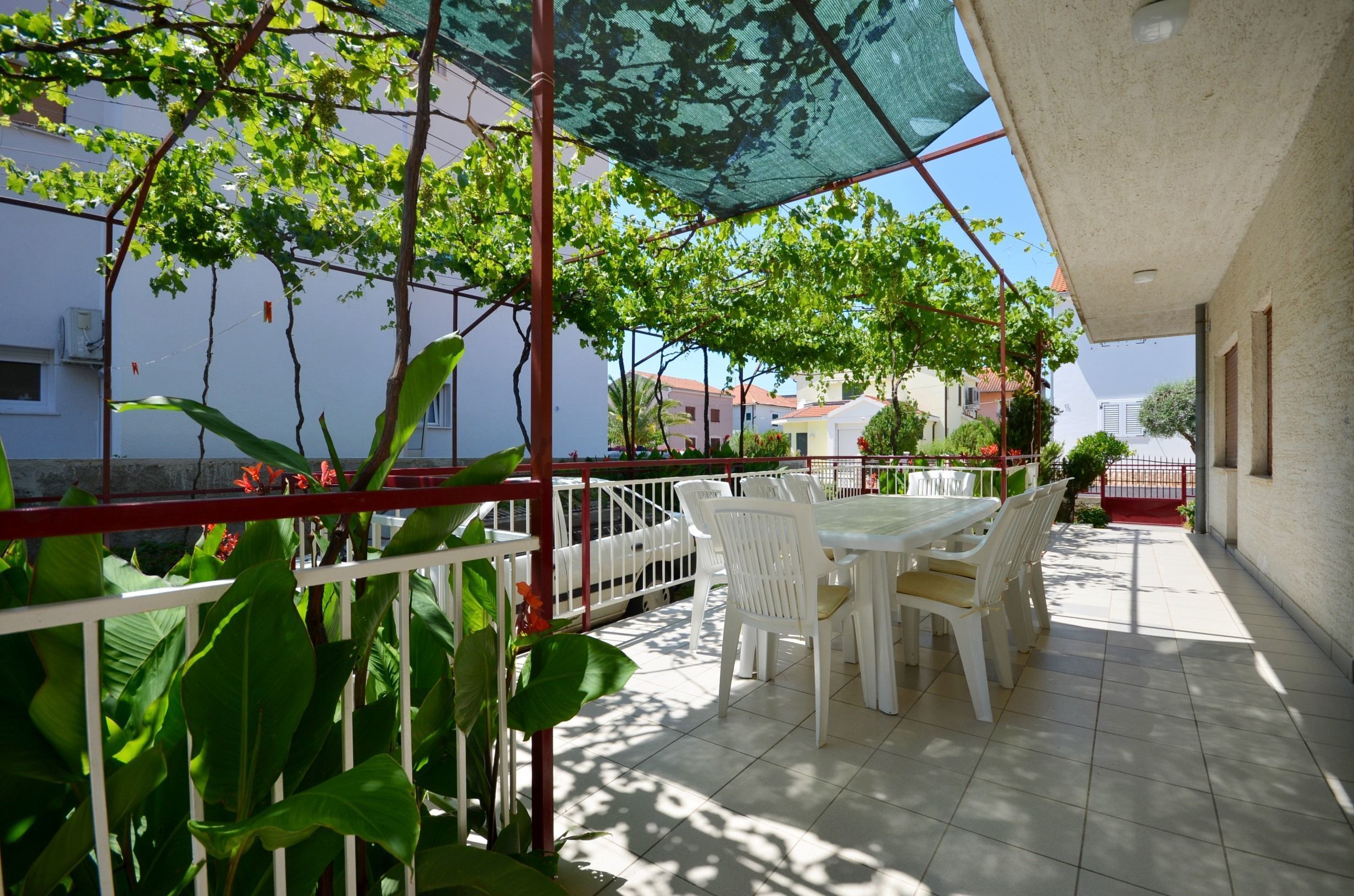 A covered outdoor patio with a table and chairs, surrounded by greenery and a view of buildings.