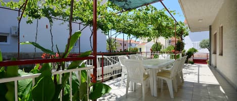 Un patio techado al aire libre con mesa y sillas, rodeado de vegetación y vistas a edificios.