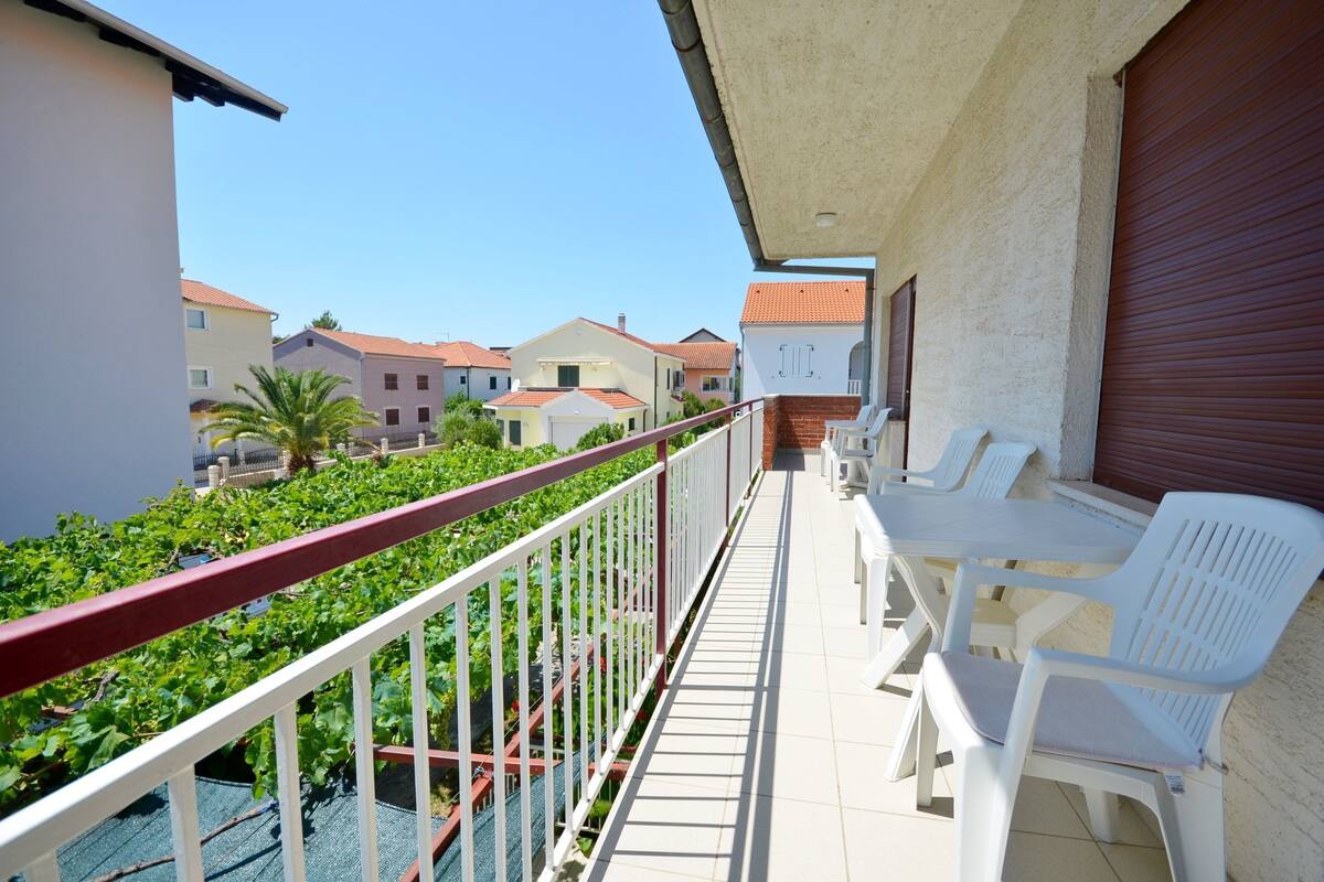 A balcony with white plastic chairs and a table, overlooking a garden with green plants and buildings in the background.