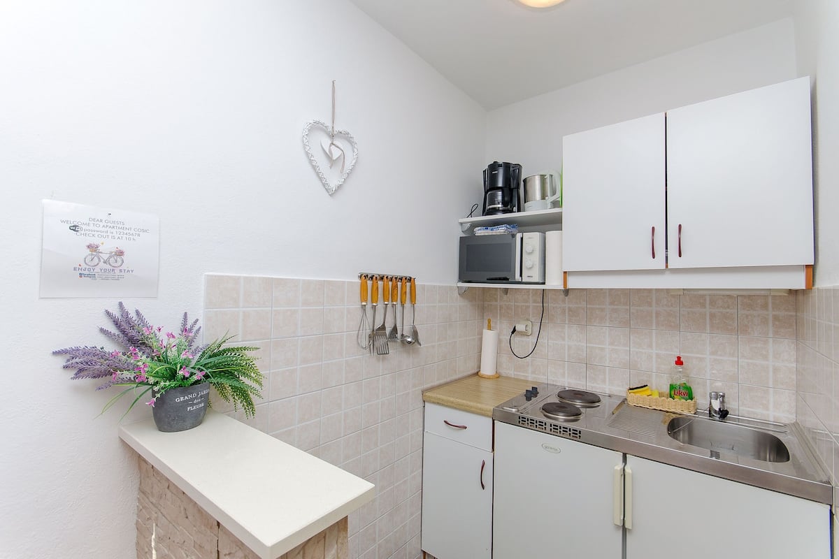 Une petite cuisine équipée avec des meubles blancs, un évier et un plan de travail. Un décor en forme de cœur orne le mur, ainsi qu’une pancarte portant un message de bienvenue.