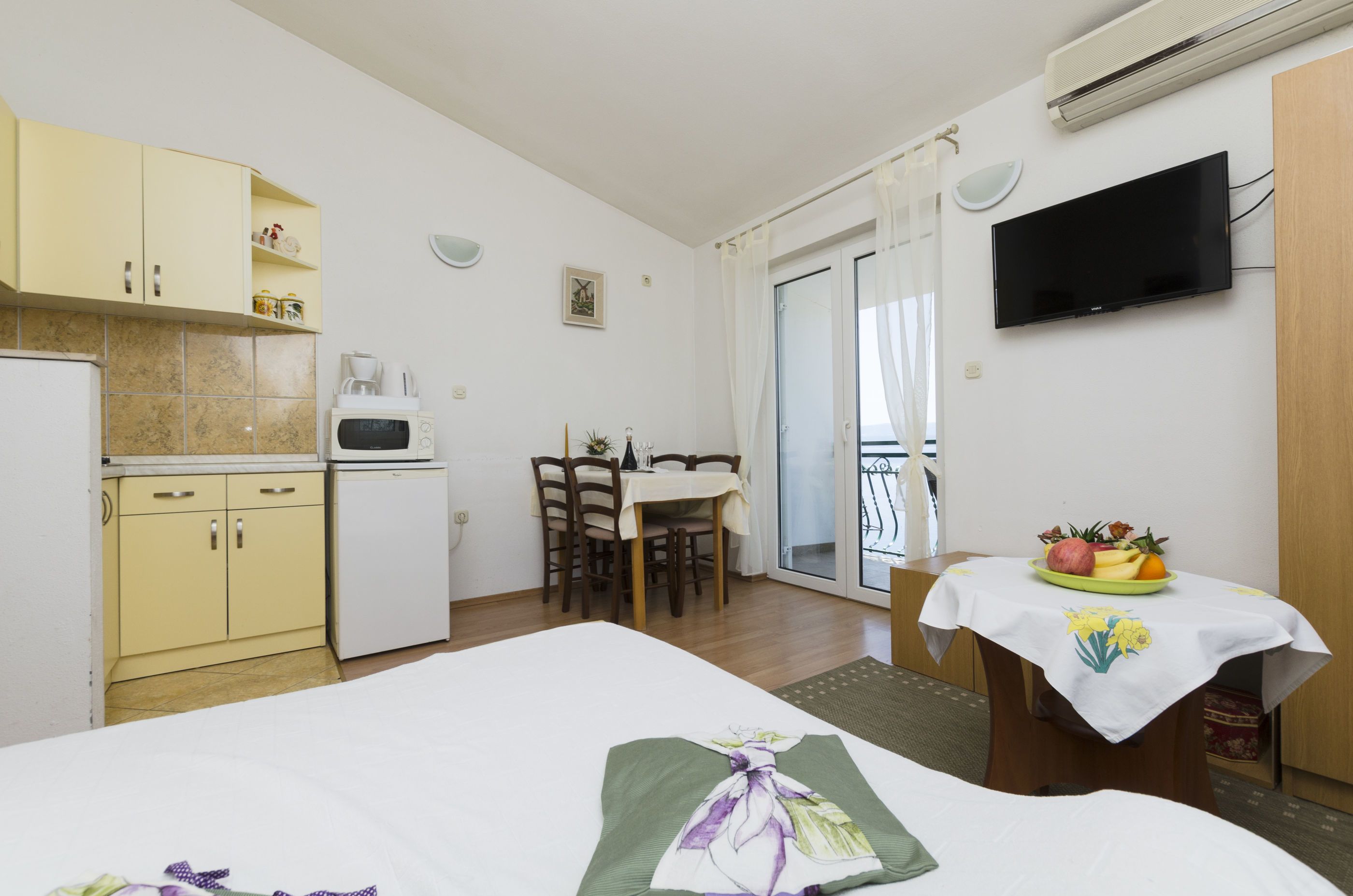 Ein Schlafzimmer mit einem Bett, einem kleinen Tisch mit Obstschale, einem Essbereich mit Stühlen, einer Küche mit Schränken und Geräten sowie einem an der Wand befestigten Fernseher.