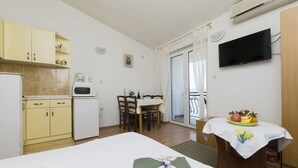 A bedroom with a bed, a small table with a fruit bowl, a dining area with chairs, a kitchen with cabinets and appliances, and a TV mounted on the wall. - Apartment Smiljana (Omis)
