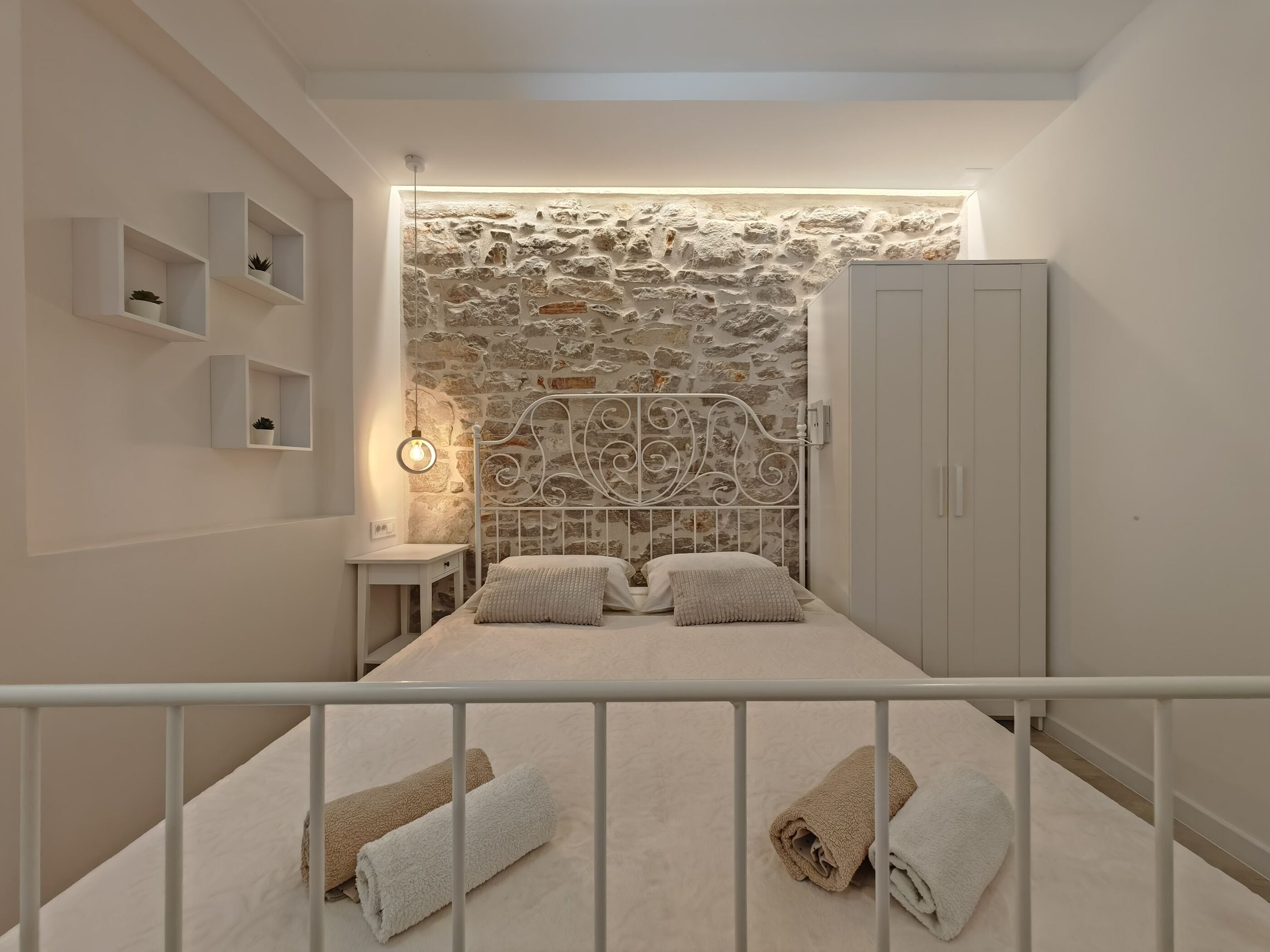 Quarto com parede de pedra, uma cama com duas almofadas, mesa de cabeceira e guarda-roupa.