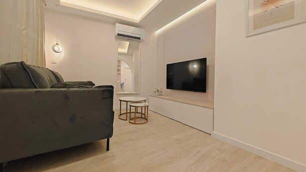 A modern living room with a grey sofa, a flat-screen TV mounted on the wall, a small round table, and a ceiling light. - Apartments Mateo (Vodice)