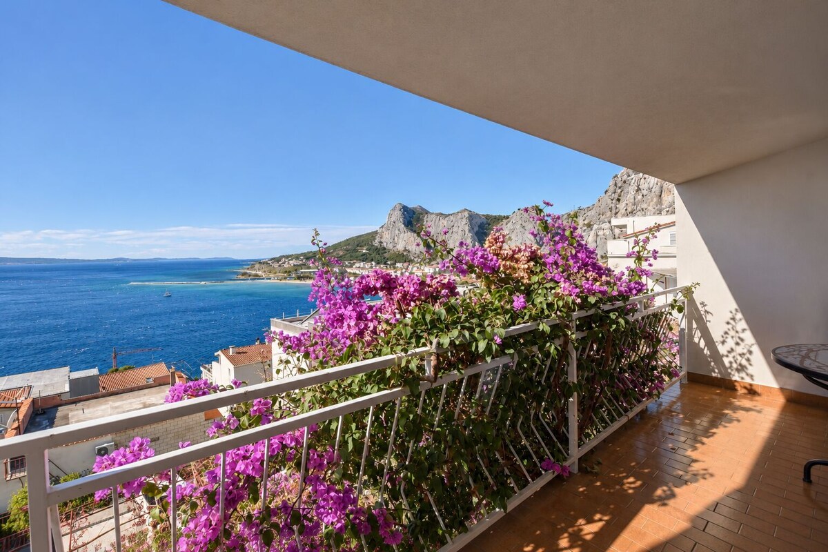 A balcony with a view of the sea and mountains, a white railing, and a wooden floor.