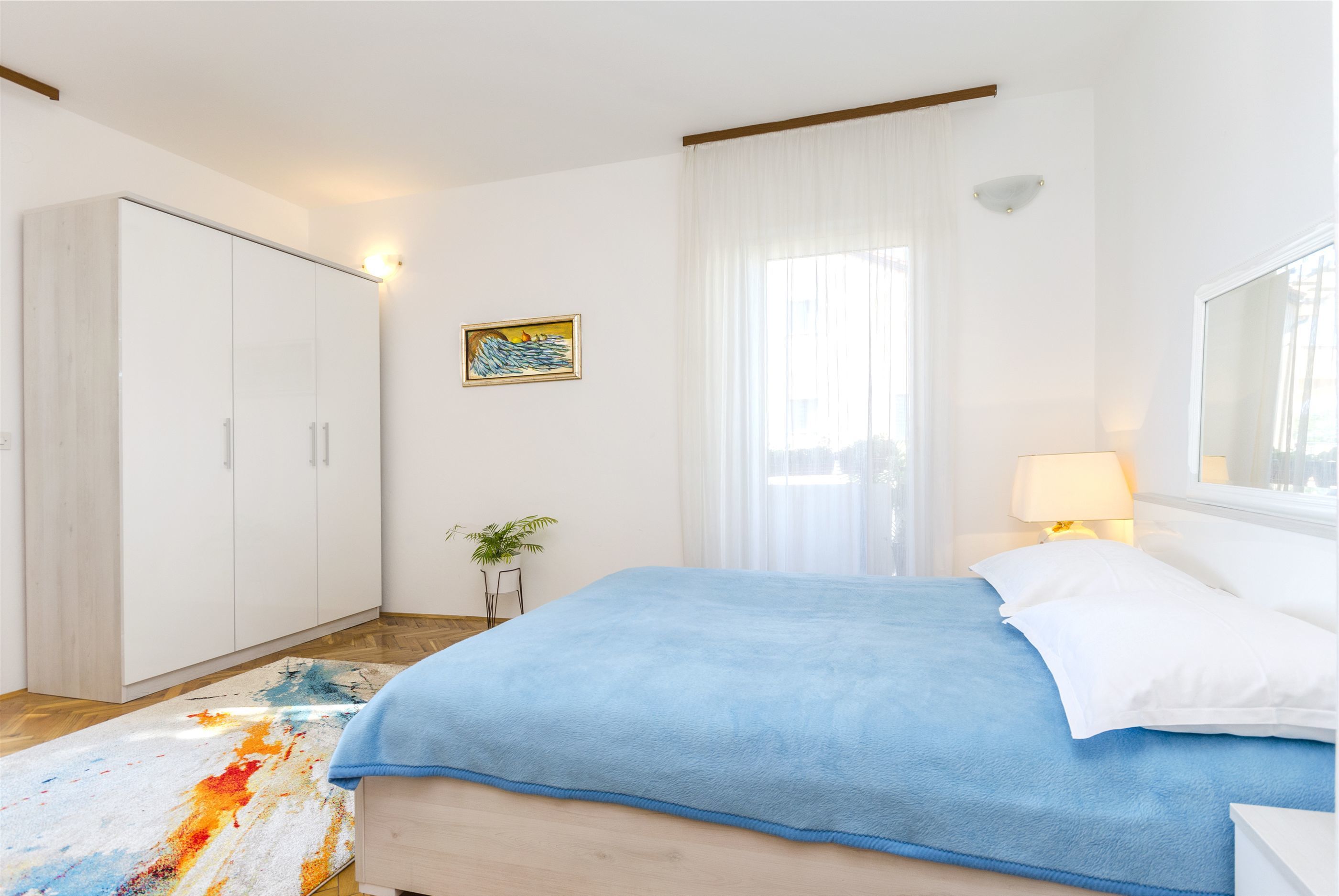 Un dormitorio con cama, armario, un cuadro, lámpara y planta.