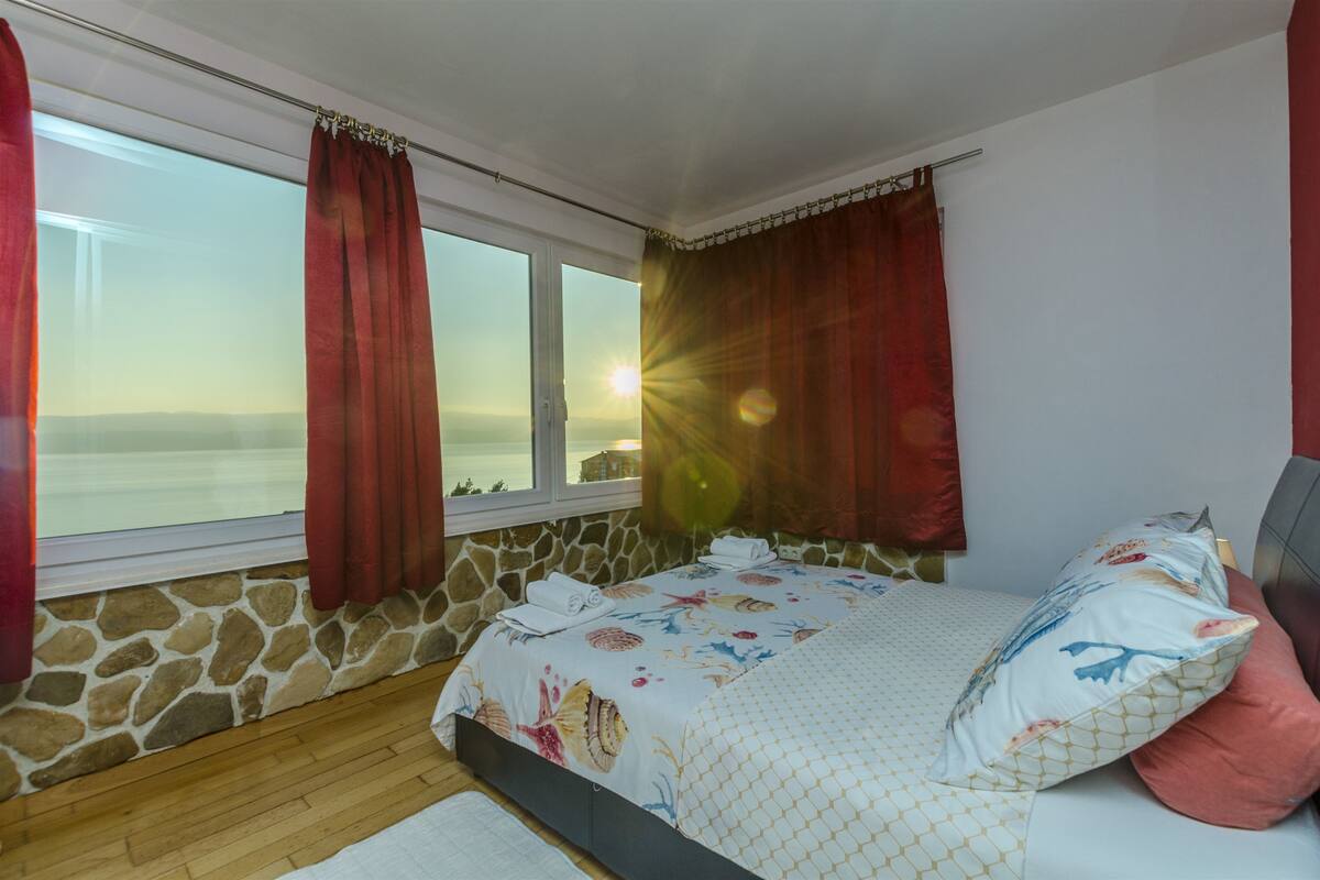 A bedroom with a stone wall, a bed with floral bedding, and a view of the sea through a window with red curtains.