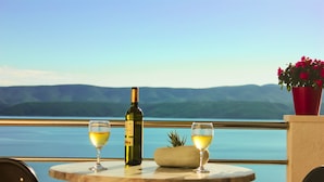 Ett bord med en vinflaska och två glas, en ananas och en röd blomkruka, med utsikt över en sjö eller ett hav och berg.